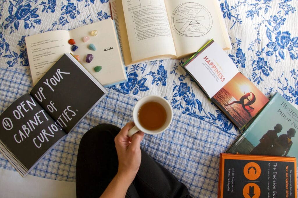 dich selbst besser kennen lernen, hier auf dem Bett sitzend, Teetasse in der Hand, umgeben von Büchern und Heilsteinen