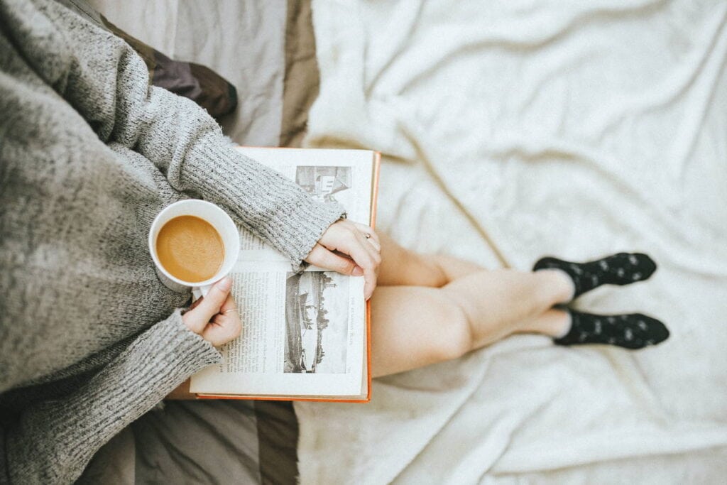 dich selbst besser kennen lernen, hier Frau mit Kaffeetasse und Buch sitzt auf Bett